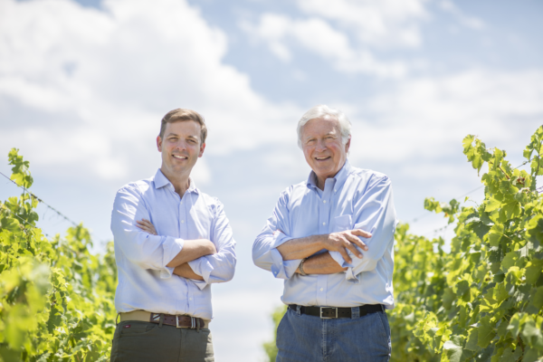 Deux hommes souriants dans les vignes du Château de Lastours, bras croisés sous un ciel bleu