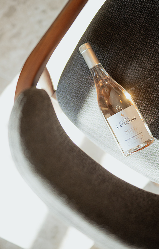 Bouteille de vin rosé du Château de Lastours posée élégamment sur un fauteuil en tissu gris