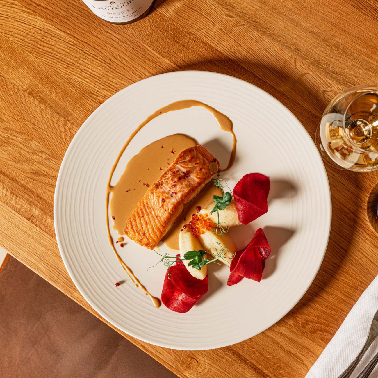 Plat gastronomique avec filet de poisson grillé, purée, légumes marinés et herbes fraîches, accompagné d’un verre de vin blanc du Château de Lastours sur table en bois.