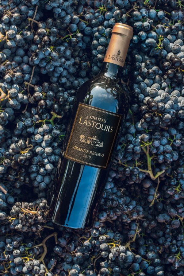 Bouteille de vin Château de Lastours Grande Réserve 2019 posée sur des grappes de raisin noir récoltées